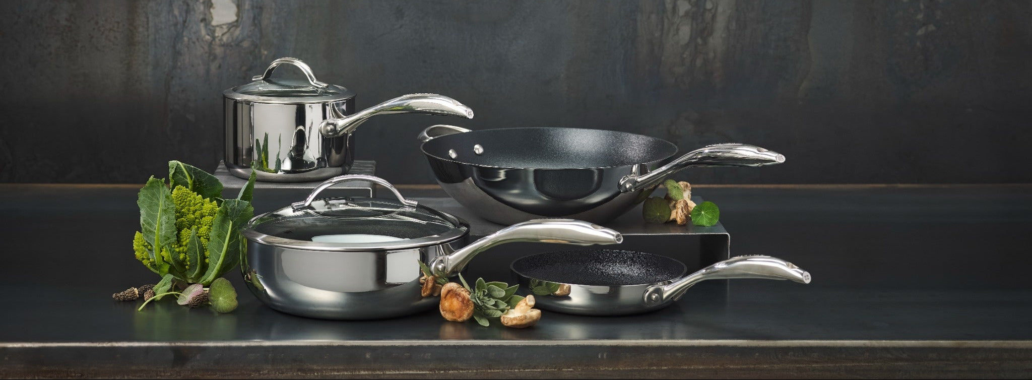 A set of Scanpan  Haptiq stainless steel and dark non-stick cookware, including a saucepan, two frying pans, and a wok, arranged on a dark surface against a textured dark gray background with green and orange vegetables as styling props.