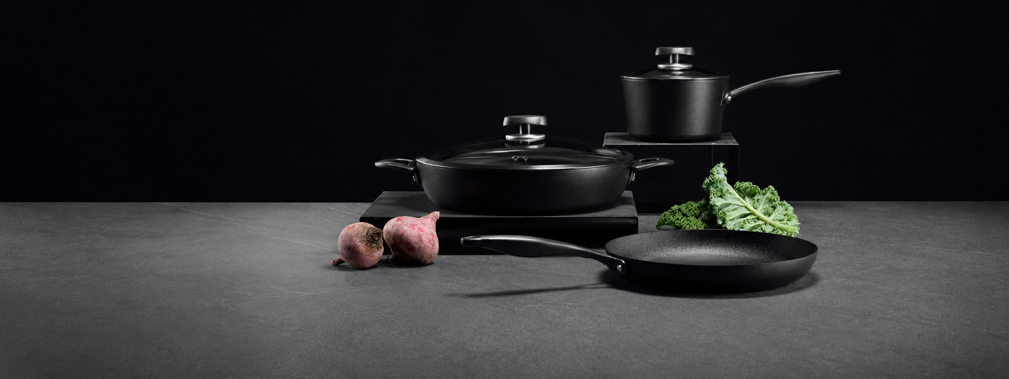 A set of  Scanpan Pro SB+ black non-stick cookware, including a saucepan, frying pan, and skillet, arranged on a dark gray surface against a black background with small food props.