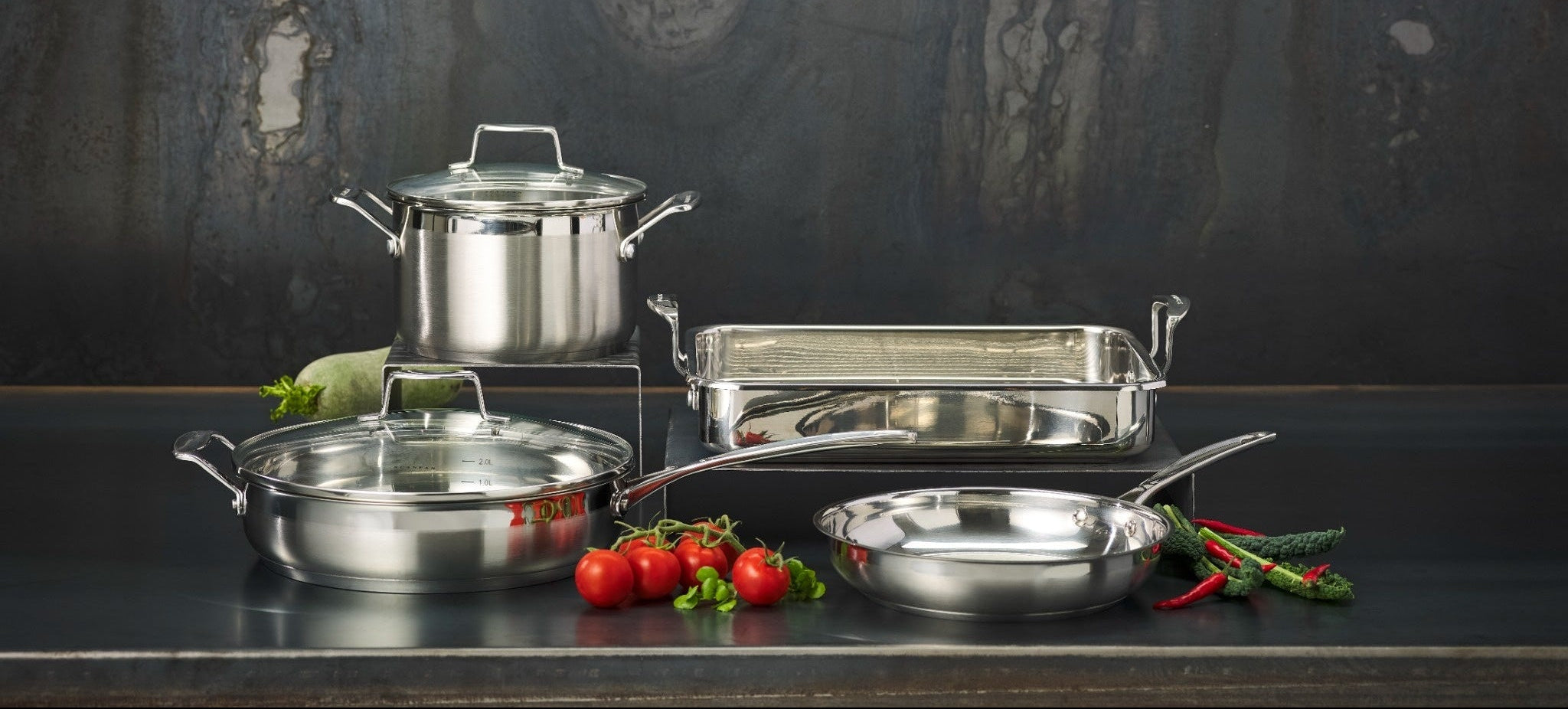 A Scanpan Impact stainless steel cookware set arranged on a dark countertop against a dark textured wall, including a pot with lid, a rectangular baking pan, a skillet, and a shallow pan, with small tomatoes and herbs as styling props.